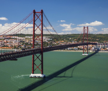 Aerial view of the 25 de Abril Bridge crossing the Tagus River between Lisbon and Almada, with its red suspension towers reflected over the water.
