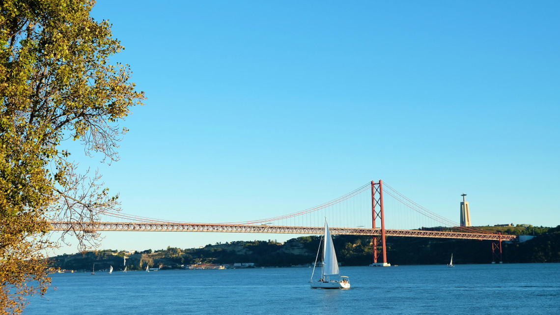 Sailboat on the Tagus River with the 25 de Abril Bridge and Christ the King statue in the background under a clear blue sky in Lisbon, Portugal