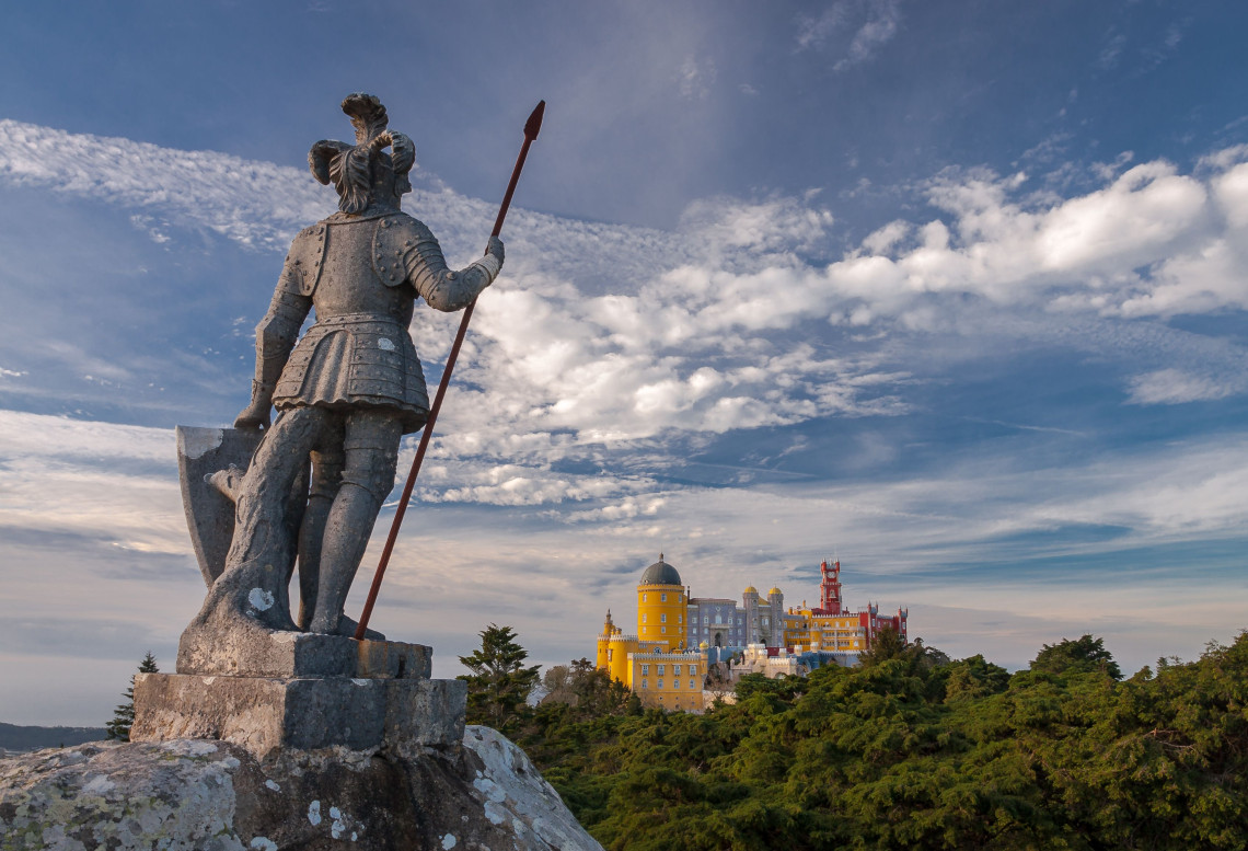 Medieval knight statue in the foreground with the colourful Pena Palace perched on a hilltop in the background under a dramatic sky in Sintra, Portugal