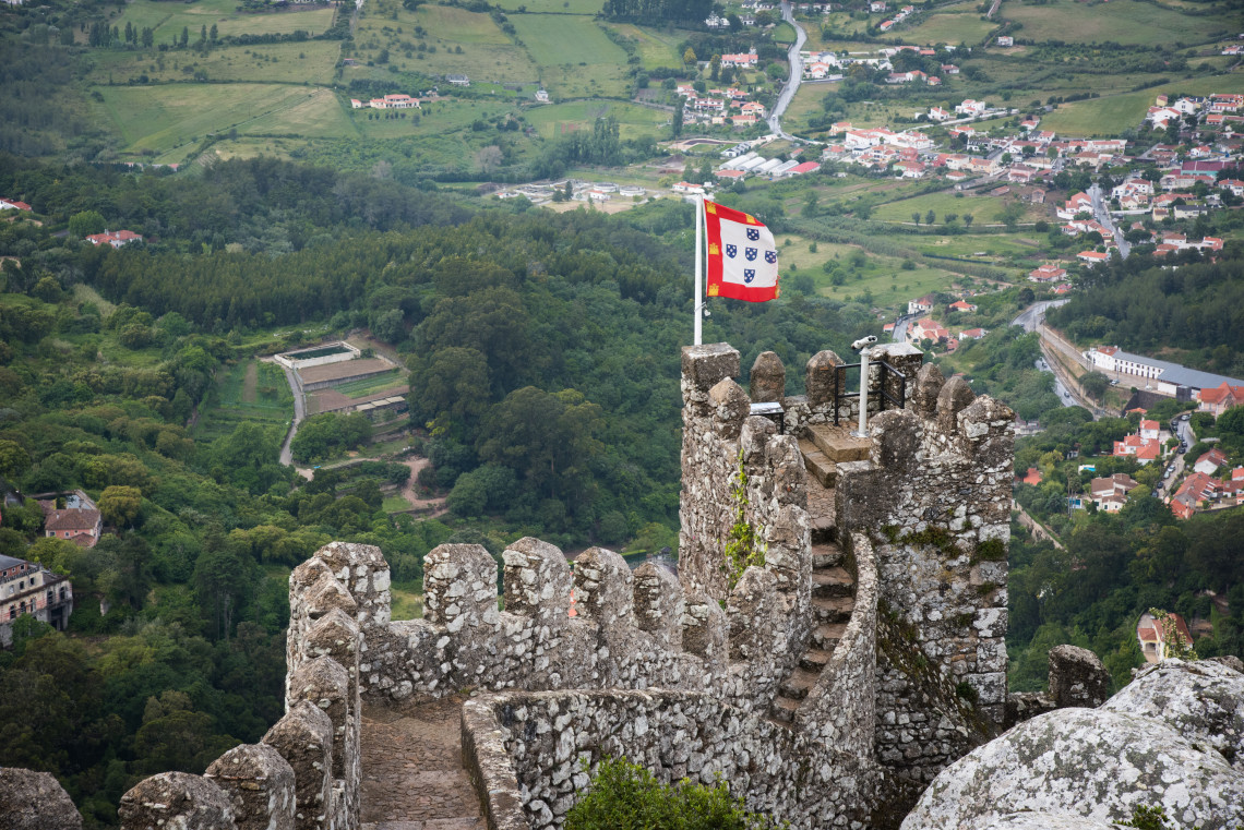 Portuguese flag flying from the ancient stone battlements of the Moorish Castle overlooking the lush green hills and village of Sintra, Portugal