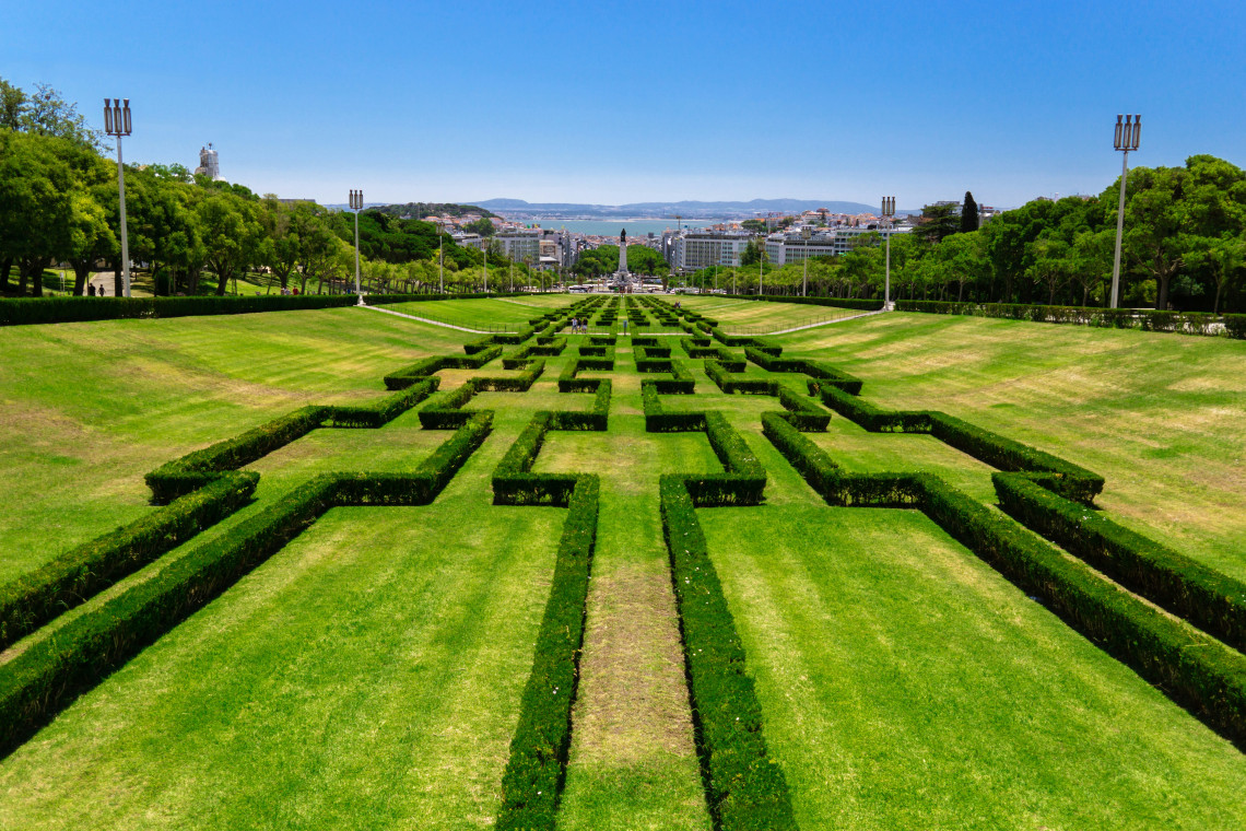 Aerial view of the manicured topiary gardens of Parque Eduardo VII stretching towards the Tagus River and Lisbon city skyline on a clear sunny day