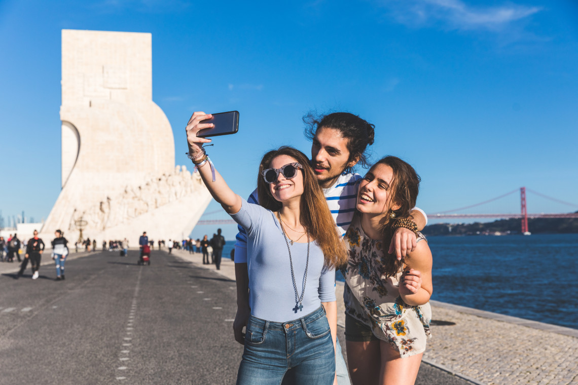 Three friends taking a selfie in front of the Monument to the Discoveries in Bel&eacute;m, Lisbon, with the 25 de Abril Bridge and Tagus River in the background on a sunny day