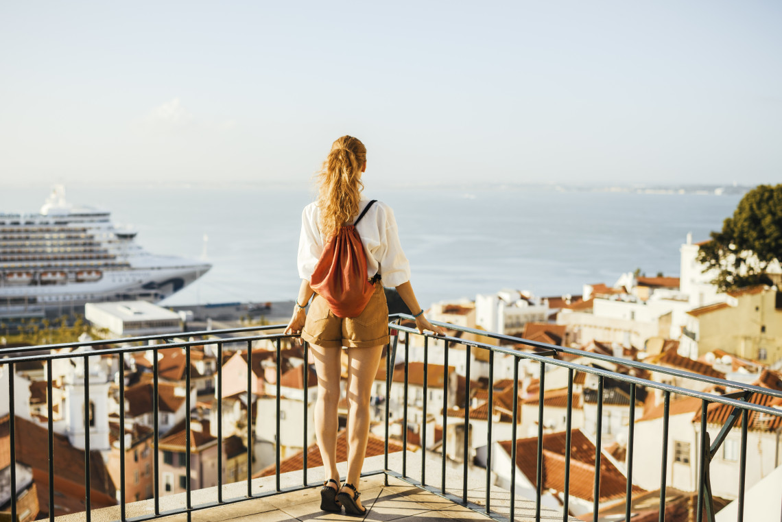 Young woman with a backpack standing on a miradouro viewpoint overlooking Lisbon's terracotta rooftops, the Tagus River and a cruise ship on a sunny day