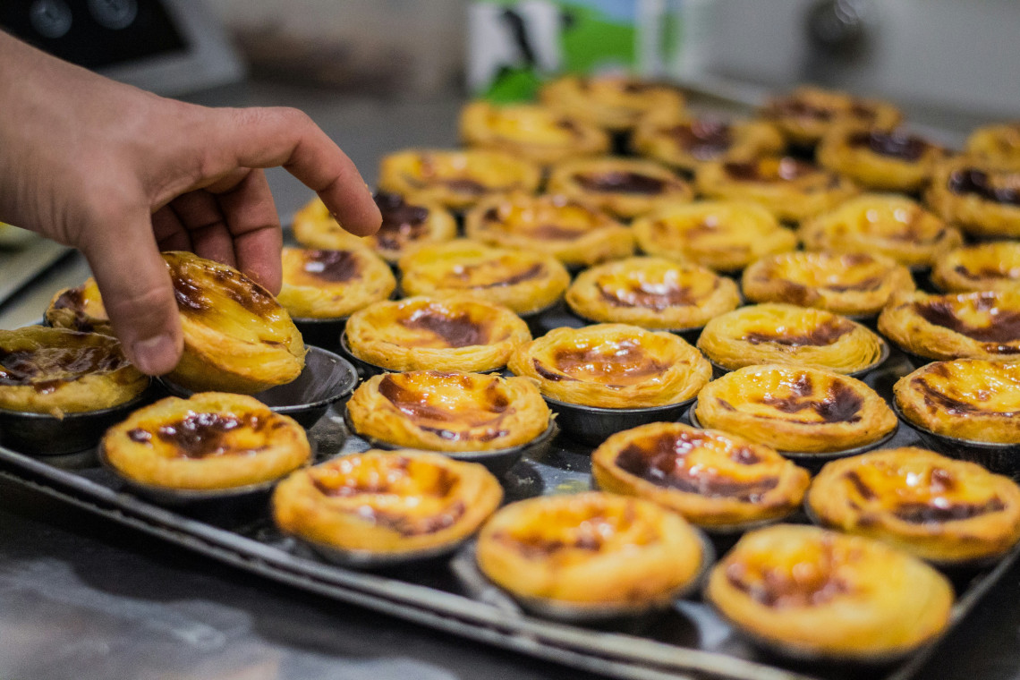 Baker's hand picking a freshly baked pastel de nata Portuguese custard tart from a tray of golden egg tarts straight out of the oven