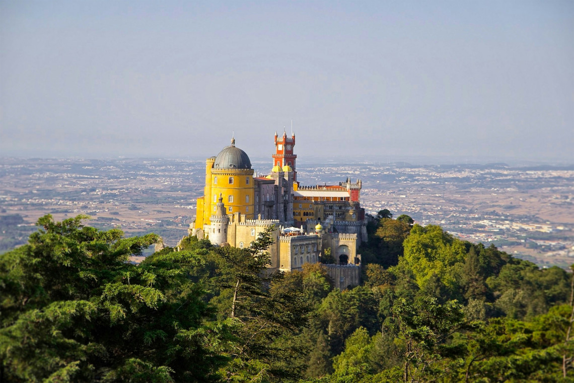 Pena Palace perched on a hilltop in Sintra, Portugal, with its vibrant yellow and red towers rising above lush green forest and panoramic countryside views