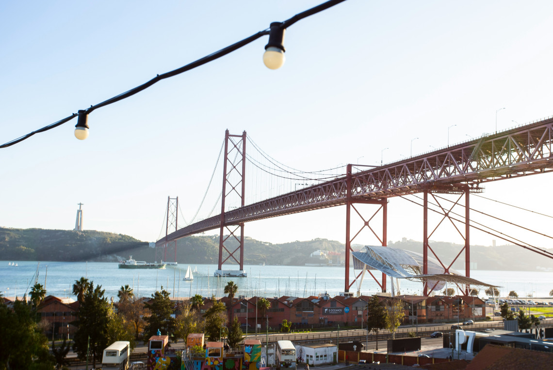 View of the 25 de Abril Bridge over the Tagus River from Alc&acirc;ntara waterfront in Lisbon, with string lights in the foreground and Christ the King statue visible in the distance