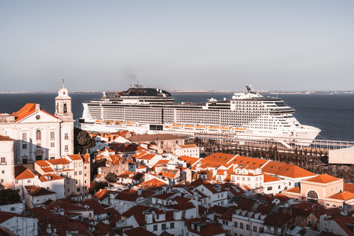 Large cruise ship docked beside Lisbon&rsquo;s historic Alfama district, with white buildings, terracotta rooftops and the Tagus River forming the waterfront landscape.