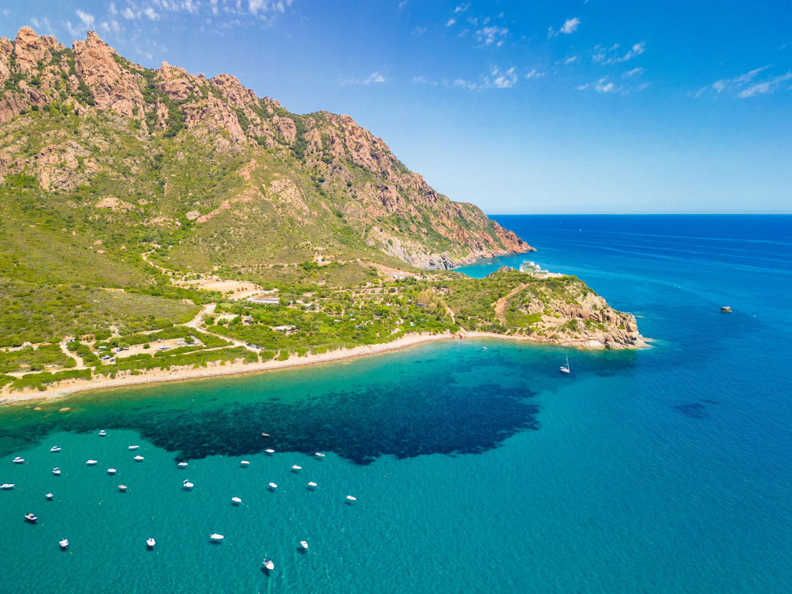 Rocky mountains descend toward clear turquoise water along the Gulf of Porto in Corsica, France, with small boats anchored near the rugged Mediterranean shoreline.