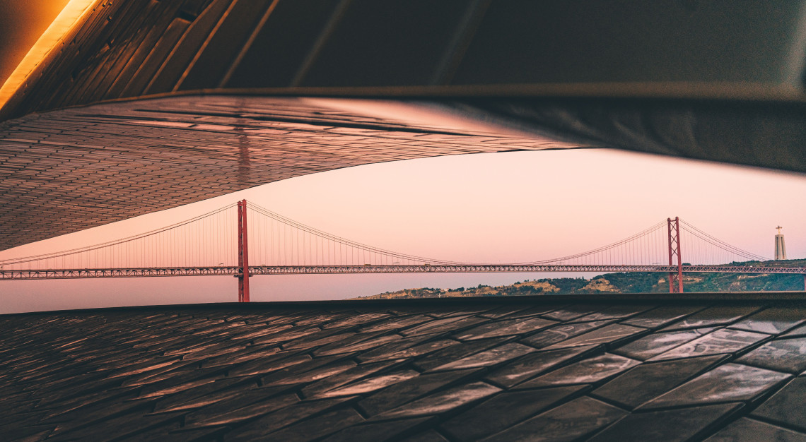 View of the 25 de Abril Bridge over the Tagus River at dusk framed by the curved roof of the MAAT museum in Lisbon, Portugal