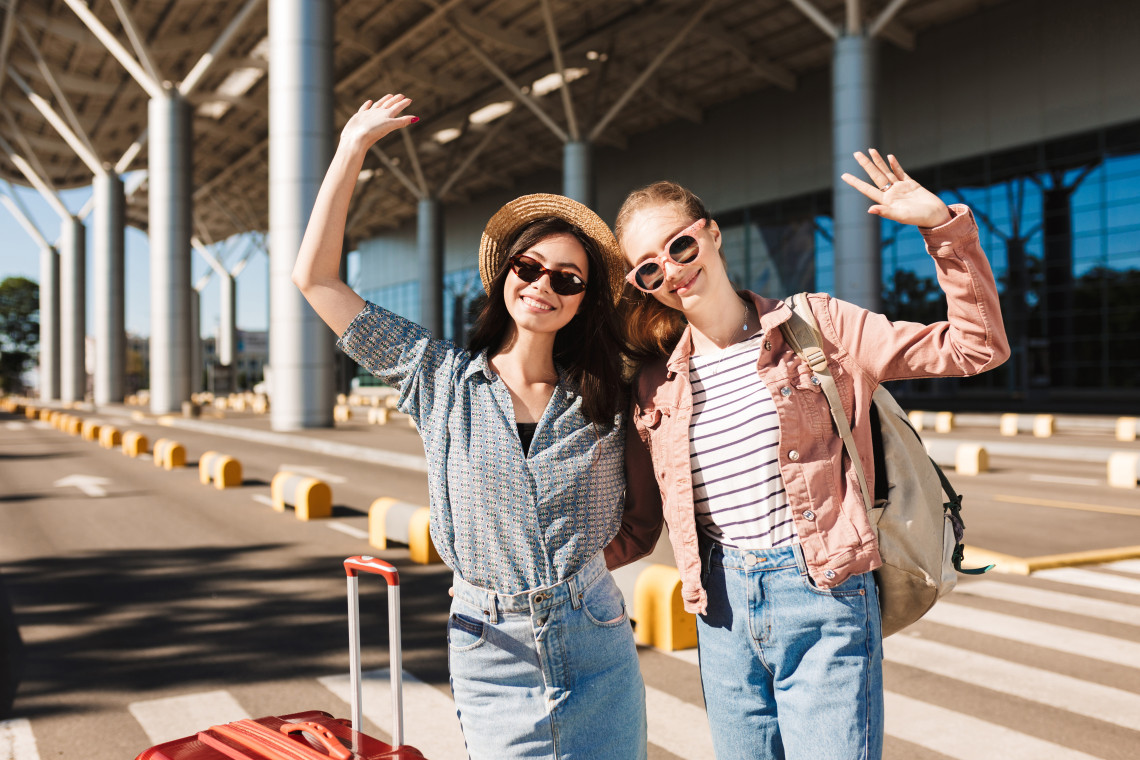 Two happy young women with sunglasses and luggage waving outside an airport terminal, excited to travel