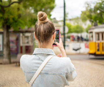 Woman photographing a classic yellow tram in Lisbon, Portugal, on a cobblestone street surrounded by trees