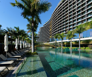Resort swimming pool lined with sun loungers and palm trees beside a large hotel building in Madeira