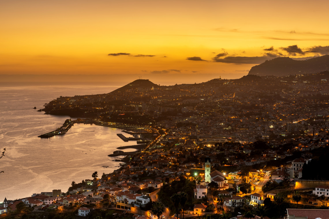 A sunset view over Funchal Bay in Funchal, Madeira Island, Portugal, with the city lights, harbor, and Atlantic coastline below. The image shows Madeira&rsquo;s coastal capital surrounded by hills and evening sky.