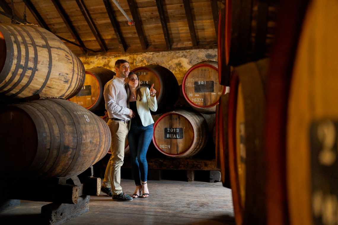 Visitors stand inside a Madeira wine cellar in Funchal, Madeira Island, Portugal, surrounded by wooden barrels used for ageing fortified wine. The image shows a traditional wine lodge setting connected to Madeira&rsquo;s historic wine culture.
