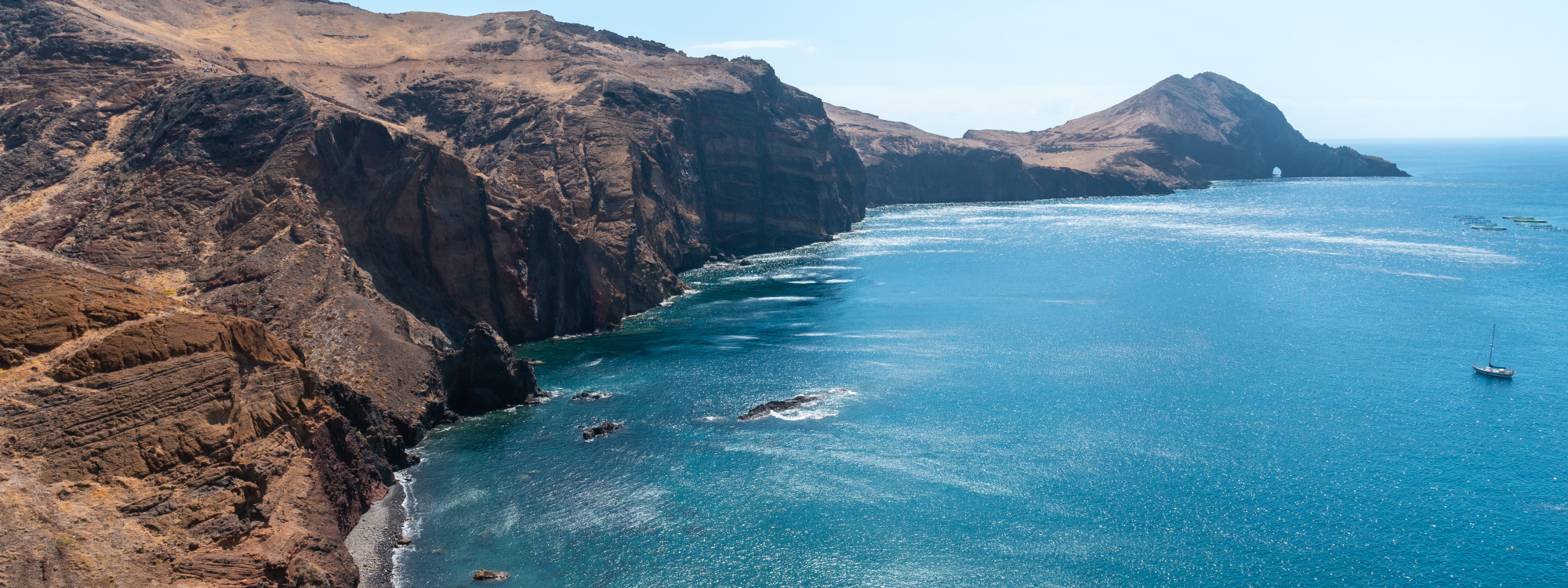 Elevated view of Ponta de São Lourenço near Caniçal, Madeira, Portugal, showing dry volcanic cliffs extending into the Atlantic Ocean. Deep blue water, rocky coves, and a small sailing boat appear along Madeira’s eastern peninsula.