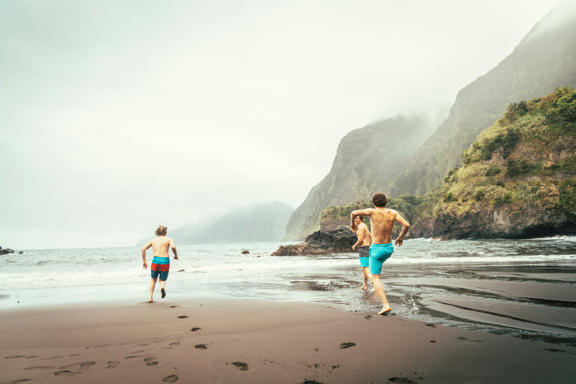 Three visitors run along the black sand of Seixal Beach in Madeira, Portugal, beside the Atlantic Ocean. Mist-covered volcanic cliffs rise above the shoreline along Madeira&rsquo;s northern coast.