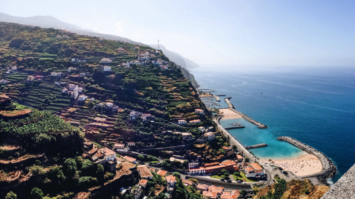 Elevated view of Calheta Beach in Madeira, Portugal, showing two sheltered golden sand bays beside the Atlantic Ocean. Terraced hillsides, houses, coastal roads, and breakwaters surround the beach on Madeira&rsquo;s southwest coast.