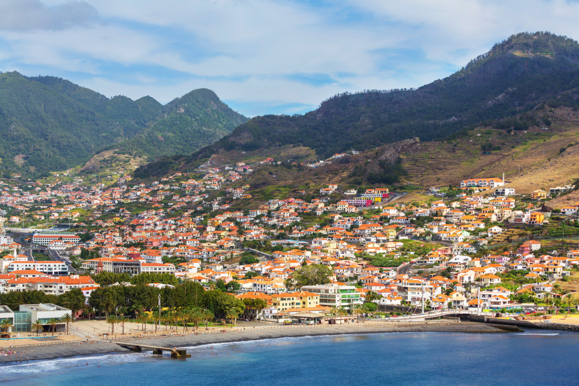 Elevated view of Machico Beach in Madeira, Portugal, with the sheltered bay and waterfront below the town&rsquo;s red-roofed buildings. Green mountain slopes rise behind Machico on Madeira&rsquo;s eastern coast.