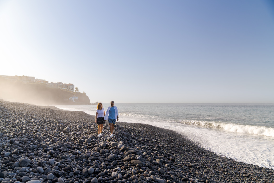 A couple walks along the dark pebble shoreline of Praia Formosa in Funchal, Madeira, Portugal. Atlantic waves reach the beach as coastal buildings and light sea mist appear in the distance.