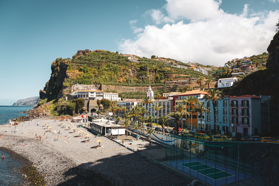 Elevated view of Praia do Vig&aacute;rio in Ponta do Sol, Madeira, Portugal, showing the pebble beach, seafront promenade, and colorful buildings beneath terraced hillsides. Visitors relax along the sheltered shoreline on Madeira&rsquo;s south coast.