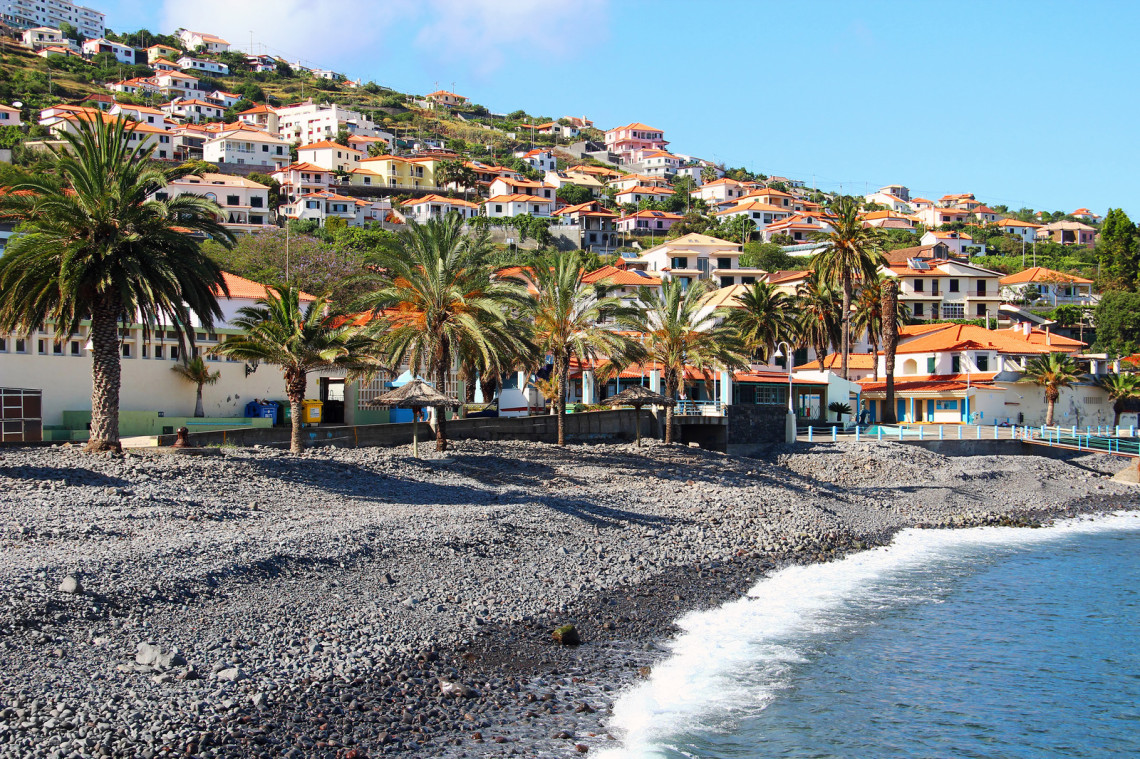 View of Santa Cruz Beach in Madeira, Portugal, showing a dark pebble shoreline, palm trees, and Atlantic waves beside the town&rsquo;s waterfront. White houses with red-tiled roofs extend across the green hillside behind the beach.