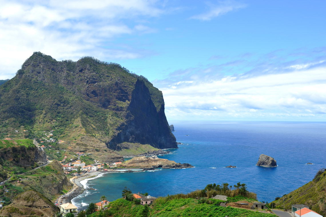 Elevated view of Alagoa Beach in Porto da Cruz, Madeira, Portugal, showing the pebble-and-sand shoreline beneath the steep volcanic slopes of Penha d&rsquo;&Aacute;guia. Rocky islets rise from the Atlantic Ocean along Madeira&rsquo;s rugged north coast.