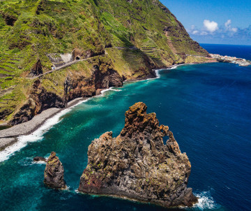 Elevated view of Penha d’Águia near Porto da Cruz, Madeira, Portugal, showing a rugged volcanic rock formation rising from the Atlantic Ocean. Steep green cliffs, a winding coastal road, and a narrow pebble shoreline frame the turquoise water.