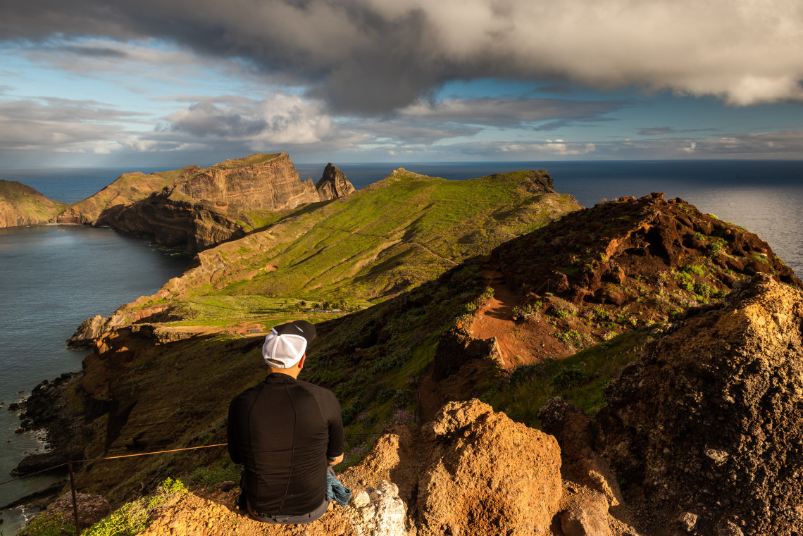 A hiker looks over Ponta de S&atilde;o Louren&ccedil;o in Cani&ccedil;al, Madeira Island, Portugal, with volcanic cliffs, green ridges, and the Atlantic Ocean in the background. The image shows the eastern peninsula&rsquo;s rugged hiking landscape and coastal views.