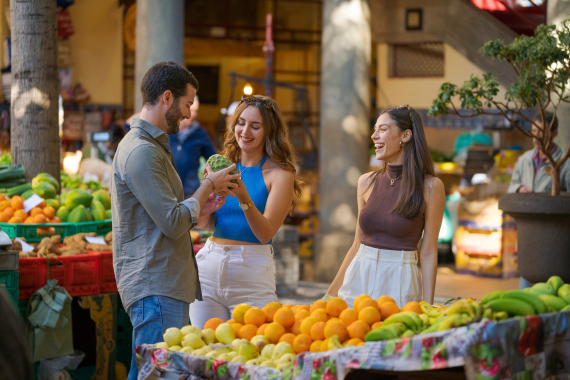 Visitors explore Mercado dos Lavradores in Funchal, Madeira Island, Portugal, surrounded by colorful fruit stands with oranges, bananas, and tropical produce. The image shows a local market experience in Madeira&rsquo;s capital city.