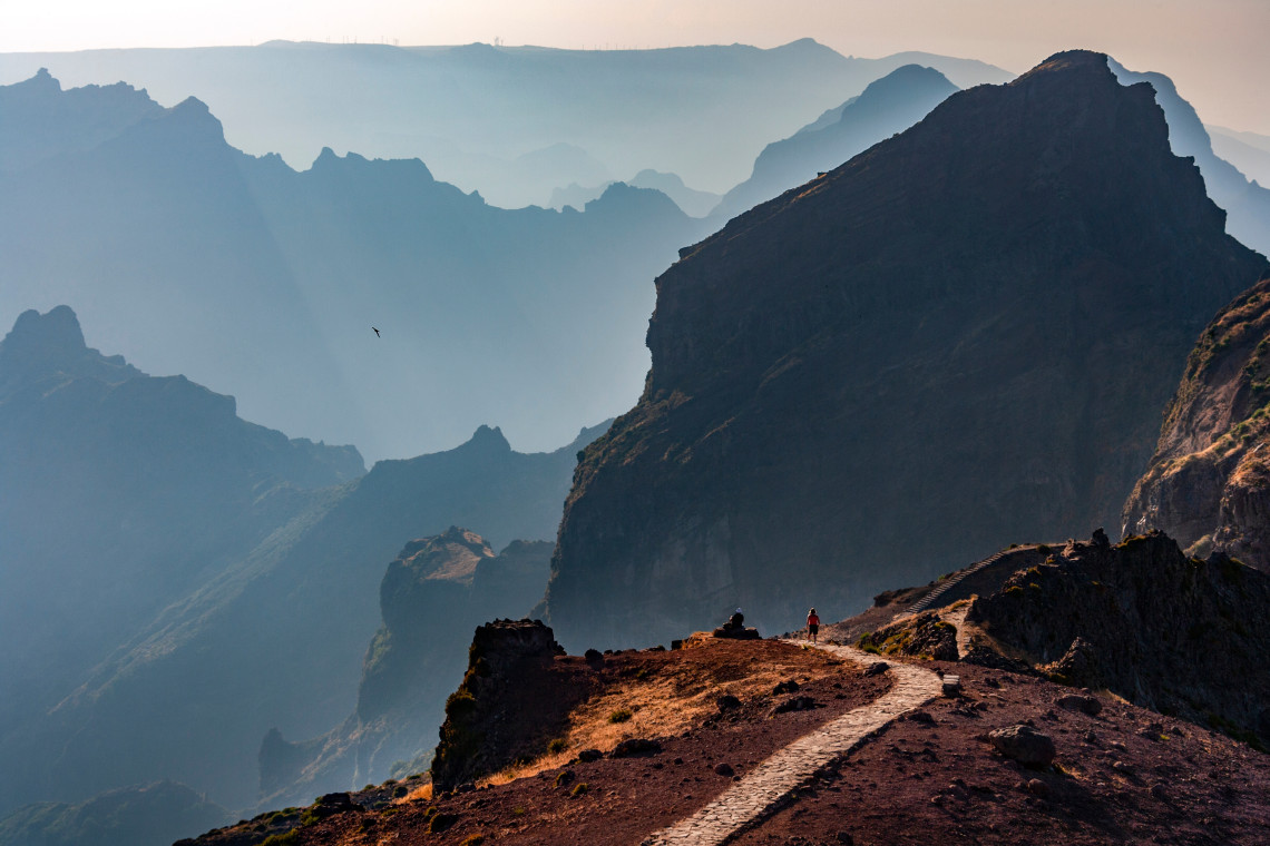 Hikers on mountain trail overlooking dramatic layered peaks, misty valleys, dark volcanic rock formations, and atmospheric haze across Madeira Island mountain landscape, Portugal