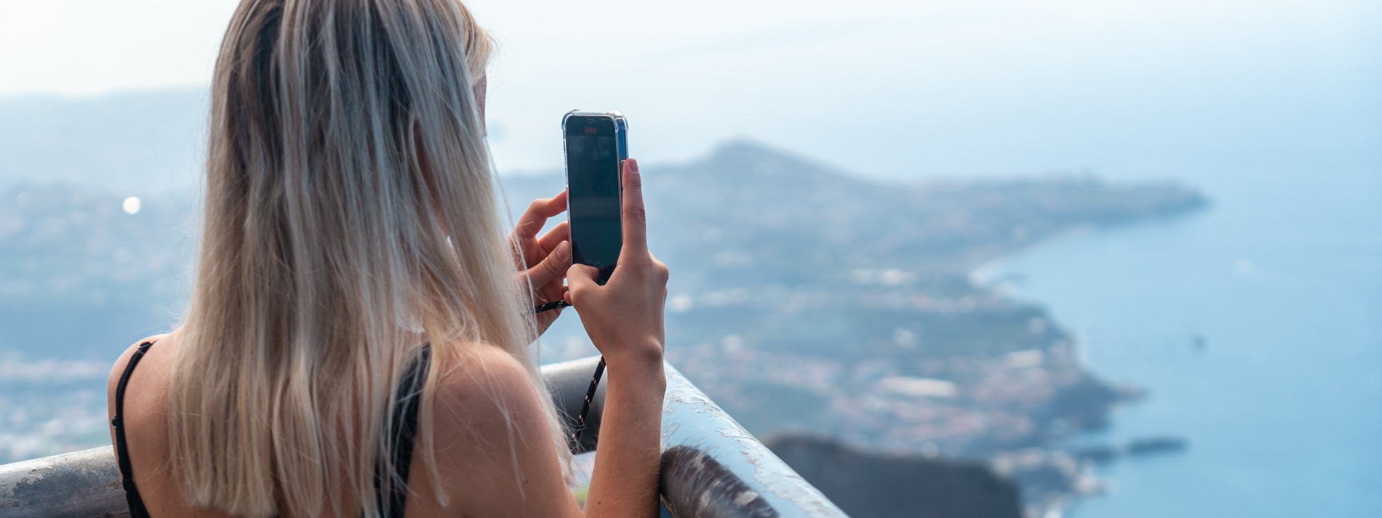 Visitor photographing the Atlantic coastline from the Cabo Girão Skywalk in Madeira, Portugal. The elevated viewpoint overlooks Câmara de Lobos, steep sea cliffs, and the island’s southern coast.