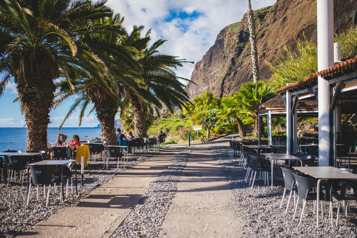 View of Faj&atilde; dos Padres in C&acirc;mara de Lobos, Madeira, Portugal, with palm trees, outdoor restaurant seating, and steep coastal cliffs beside the Atlantic Ocean. The image shows a peaceful seaside setting on Madeira&rsquo;s south coast.