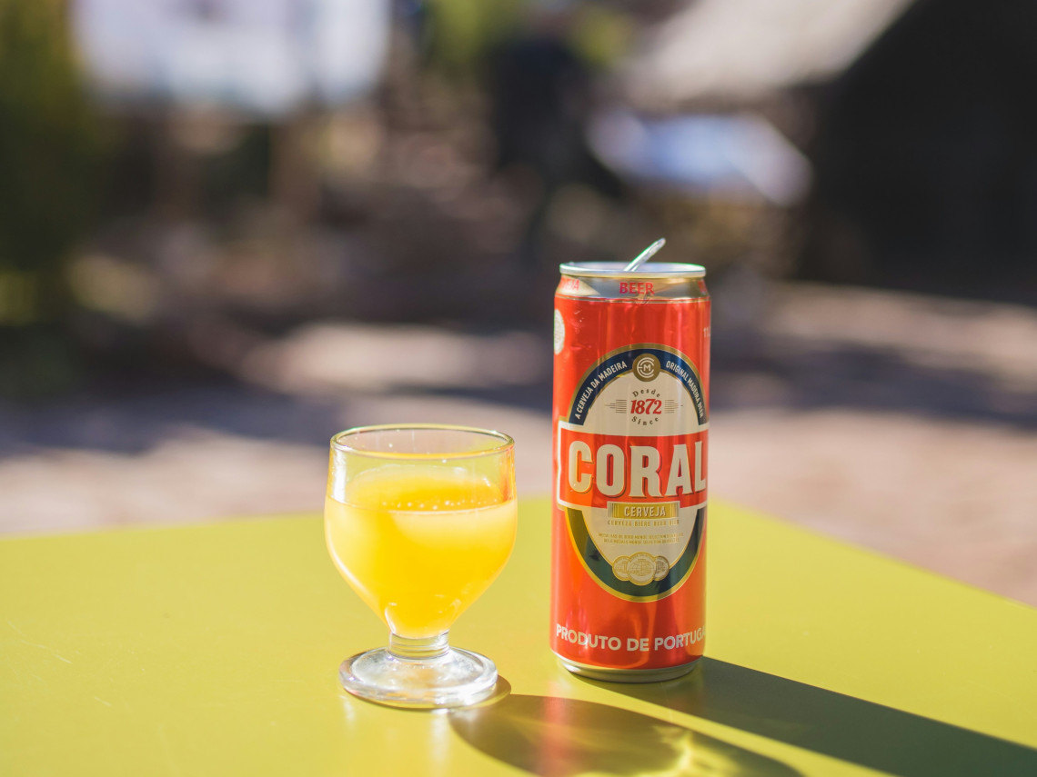 A can of Coral beer and a glass of orange juice sit on an outdoor terrace table in Madeira, Portugal. The image highlights local drinks and relaxed island dining in a sunny setting.