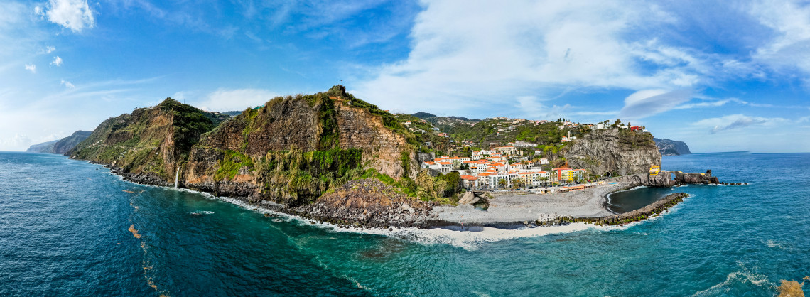 Panoramic view of Ponta do Sol in Madeira, Portugal, showing the coastal village, pebble beach, steep volcanic cliffs, and Atlantic Ocean. The image highlights Madeira&rsquo;s south coast landscape with dramatic cliffs and seaside architecture.