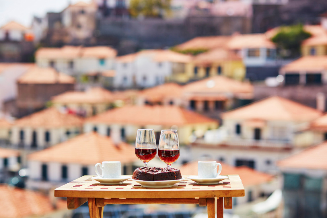 Bolo de Mel from Madeira Island, Portugal, served with coffee and glasses of Madeira wine overlooking traditional terracotta rooftops. The image shows a classic Madeiran sweet connected to the island’s dessert and wine culture.
