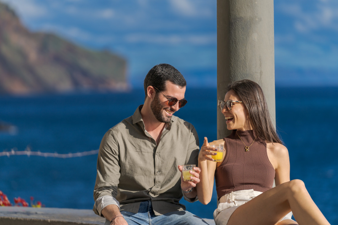 Visitors enjoy poncha, a traditional drink from Madeira Island, Portugal, with Atlantic Ocean and coastal cliffs in the background. The image shows a relaxed Madeiran food and drink experience overlooking the island’s coastline.