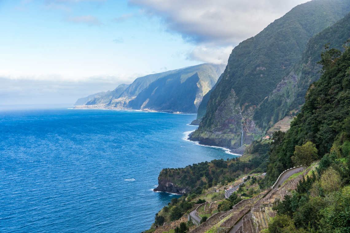 A scenic view of Madeira Island&rsquo;s north coast in Portugal, with steep green cliffs, winding coastal roads, and the Atlantic Ocean below. The image shows Madeira&rsquo;s mountainous shoreline and rugged seaside landscape.
