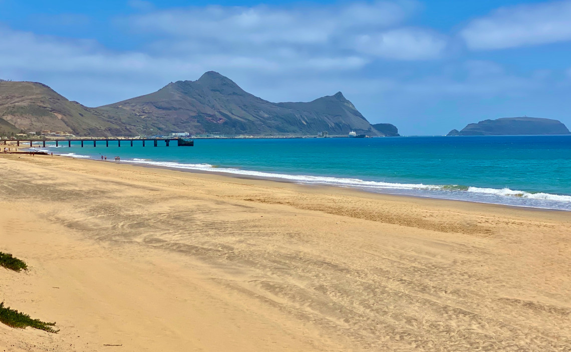 Porto Santo Beach in Porto Santo, Portugal, with golden sand, turquoise Atlantic water, and mountain scenery along the island&rsquo;s coastline. The image shows the beach and pier with views toward Porto Santo&rsquo;s rugged eastern landscape.