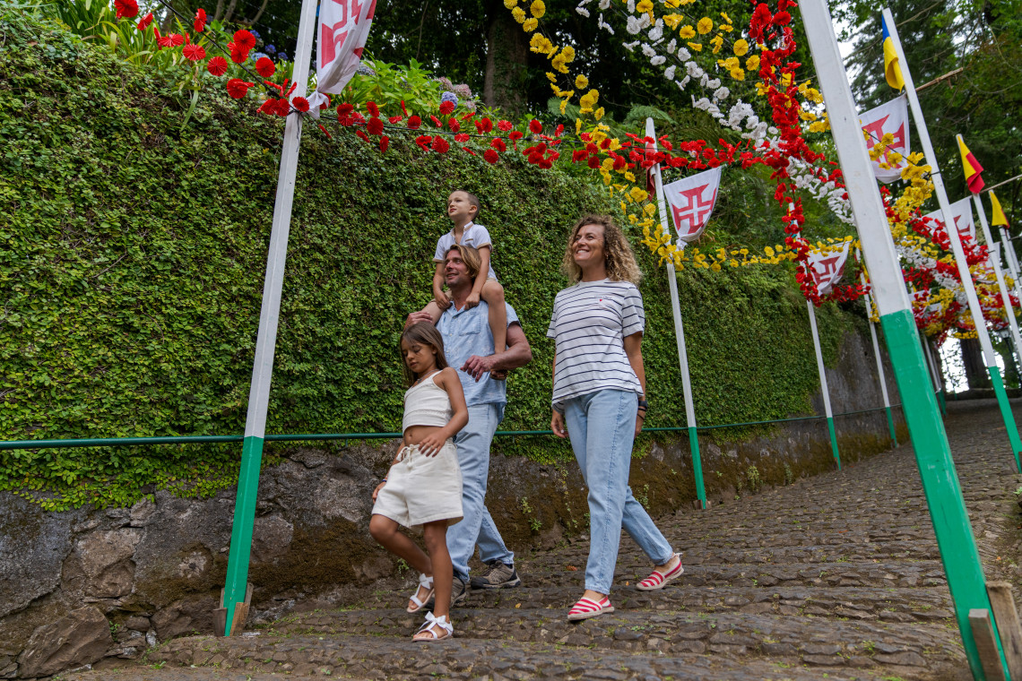 A family walks through a decorated street during a Madeira arraial on Madeira Island, Portugal, surrounded by floral garlands, festival flags, and cobblestone paths. The image shows a traditional Madeiran community celebration with festive street decorations.