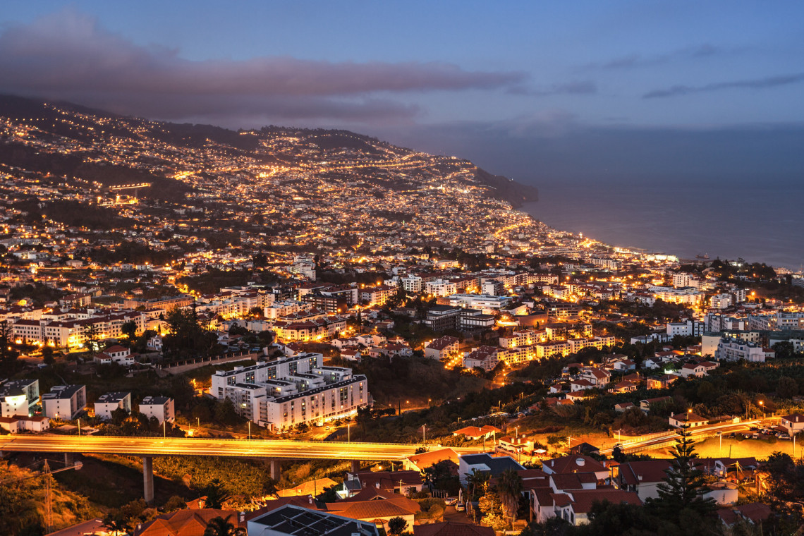 Elevated dusk view over Funchal, Madeira, Portugal, showing illuminated neighborhoods extending across the hills toward the Atlantic Ocean. Roads, residential buildings, and the coastal city center are visible beneath low clouds.