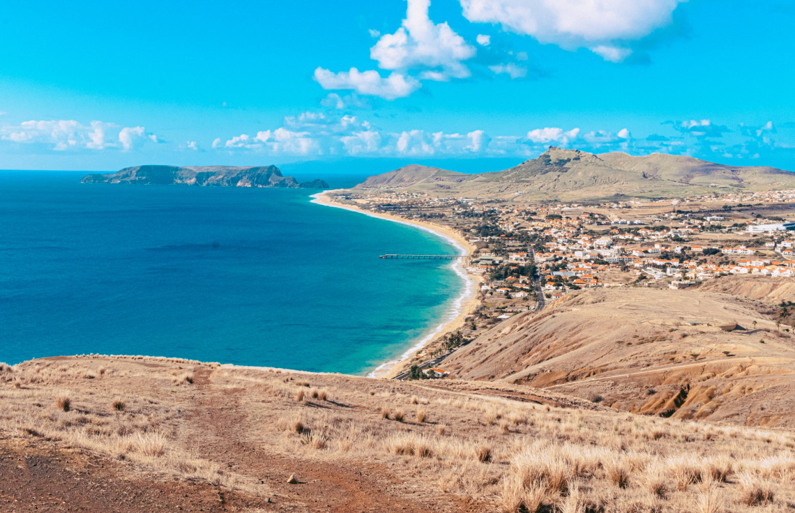 Aerial view of Porto Santo Island with pristine sandy beach, turquoise Atlantic waters, arid volcanic landscape, small town, and neighboring islands under clear blue sky, Madeira archipelago