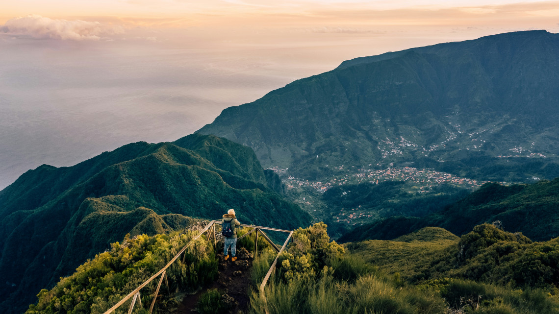 Hiker standing on wooden railing at mountain viewpoint overlooking lush green peaks, deep valleys, and town settlement nestled below during golden hour in Madeira, Portugal