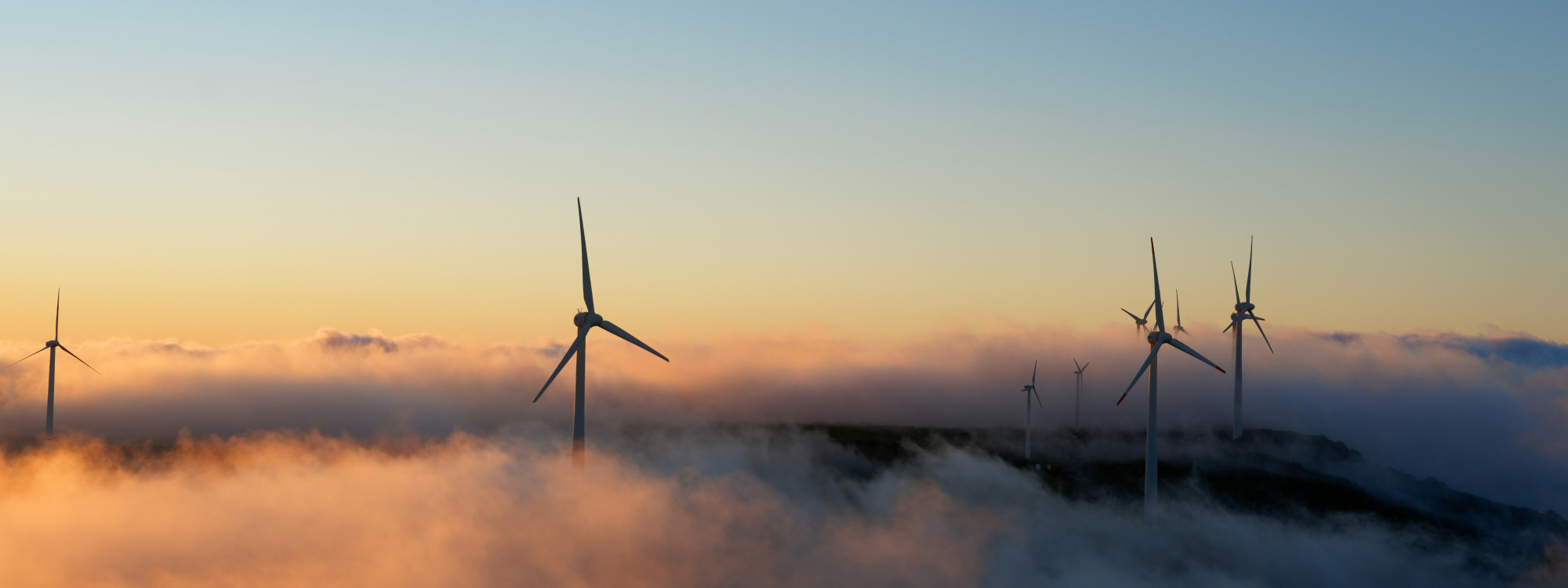 Wind turbines rise above low clouds at Paul da Serra in Madeira, Portugal, during sunset. The image shows the island’s high-altitude plateau surrounded by mist and warm evening light.