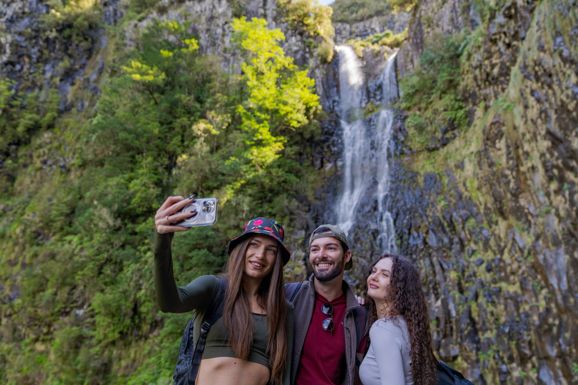 Three travelers take a selfie in front of Risco Waterfall in Raba&ccedil;al, Madeira, Portugal. The tall cascade flows over dark volcanic rock surrounded by dense Laurisilva vegetation along the Levada do Risco hiking route.