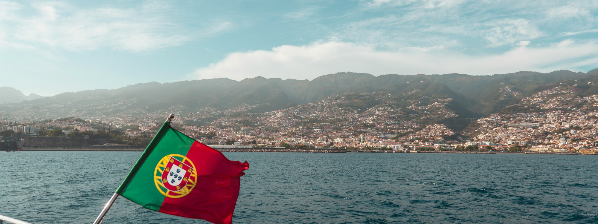 Portuguese flag flying from a boat in Funchal Bay, Madeira, with the densely built hillsides and mountainous landscape of Funchal rising behind the Atlantic waterfront.