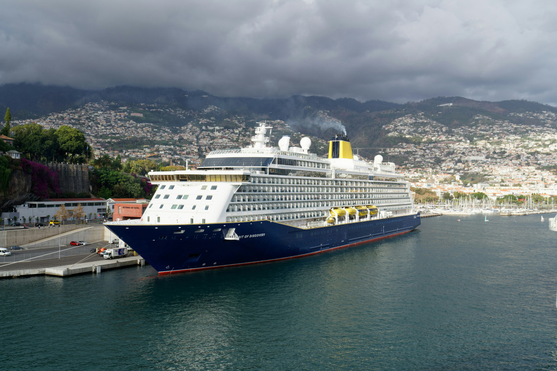 Large cruise ship docked at the Port of Funchal on Madeira Island, Portugal, with the hillside city and green mountains forming the backdrop.