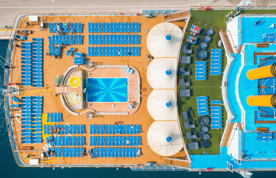 Top-down view of a cruise ship&rsquo;s open-air pool deck, with rows of sun loungers, swimming pools and shaded relaxation areas arranged across the upper deck.