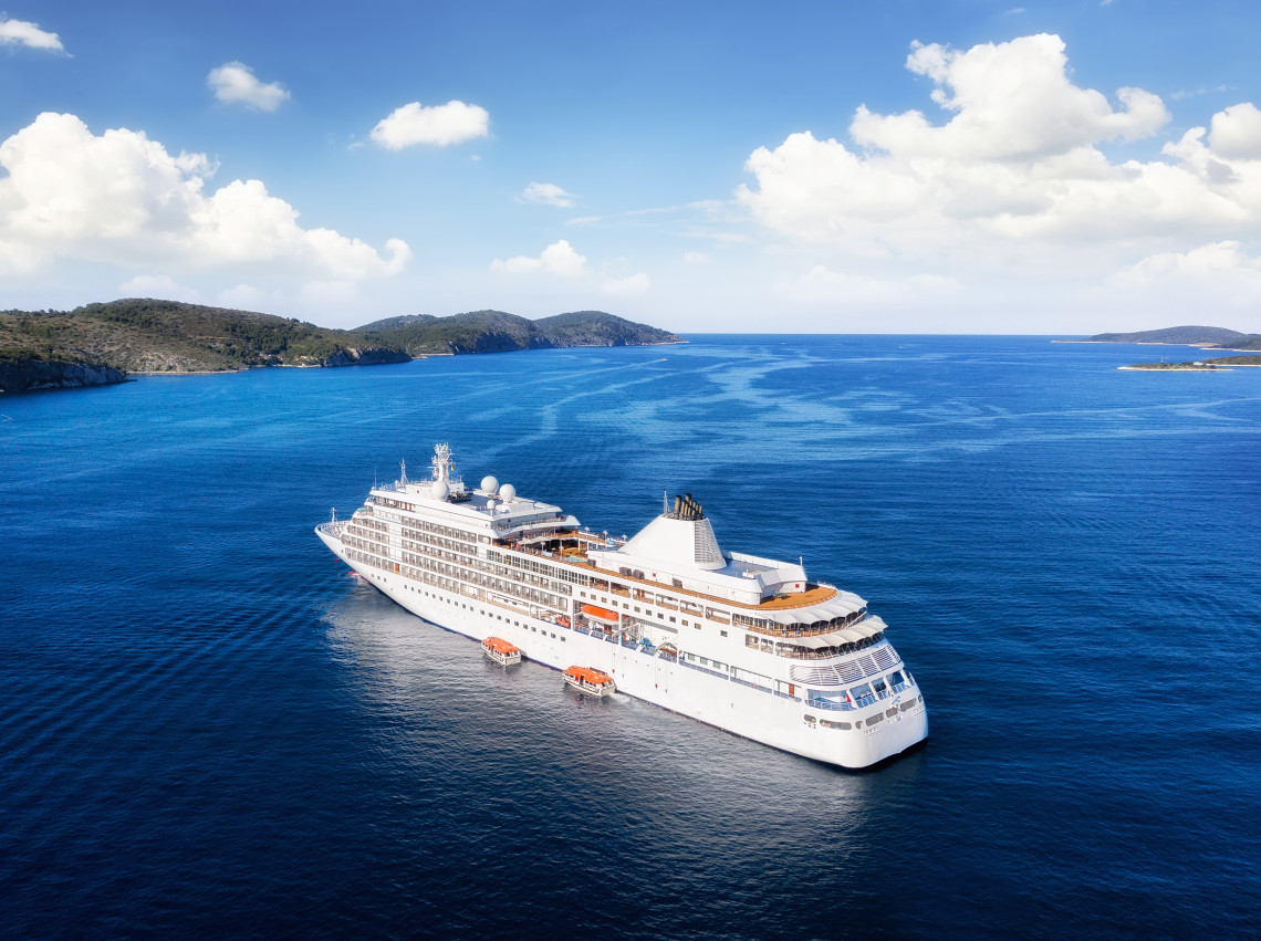 White cruise ship sailing through deep blue Mediterranean waters between low rocky islands beneath a bright sky, photographed from an elevated aerial perspective.