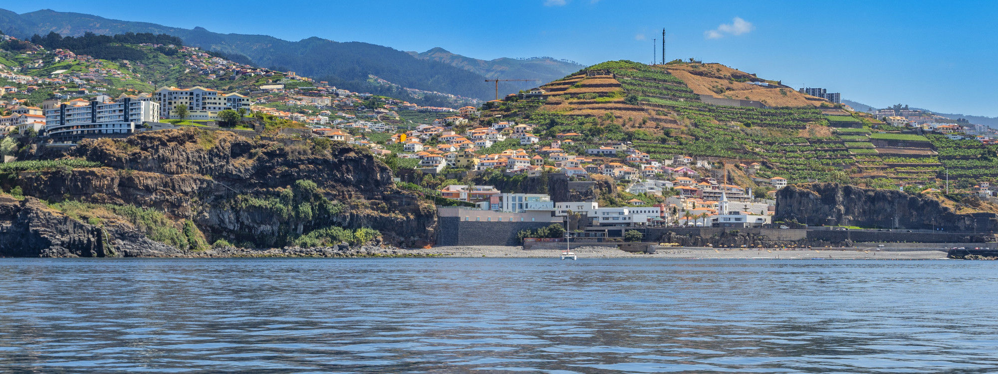 View from the Atlantic Ocean toward Praia Formosa in Funchal, Madeira, Portugal. The coastline features dark volcanic cliffs, a waterfront promenade and buildings extending across cultivated hillsides.