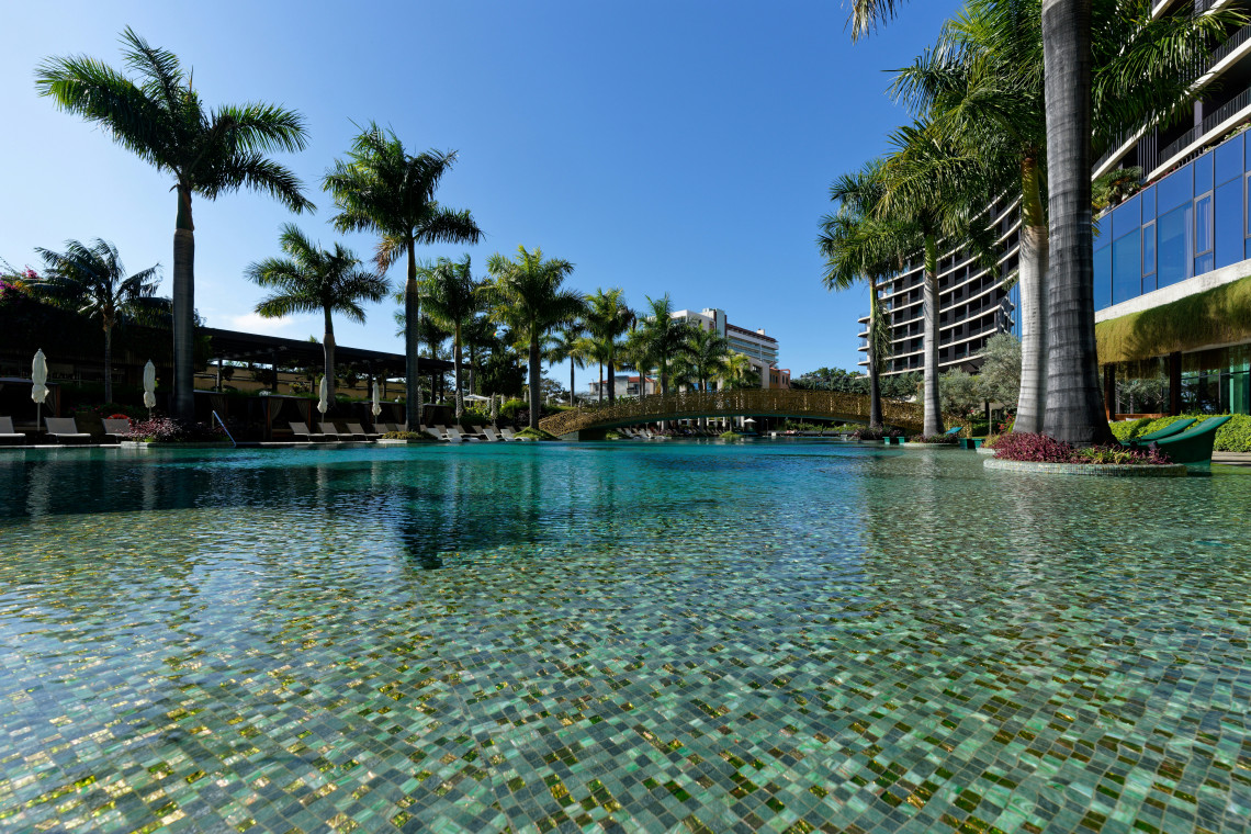 The outdoor lagoon-style pool at Savoy Palace in Funchal, Madeira, Portugal, is surrounded by palm trees, landscaped gardens and contemporary hotel buildings. A decorative bridge crosses the water beside submerged loungers and poolside seating.